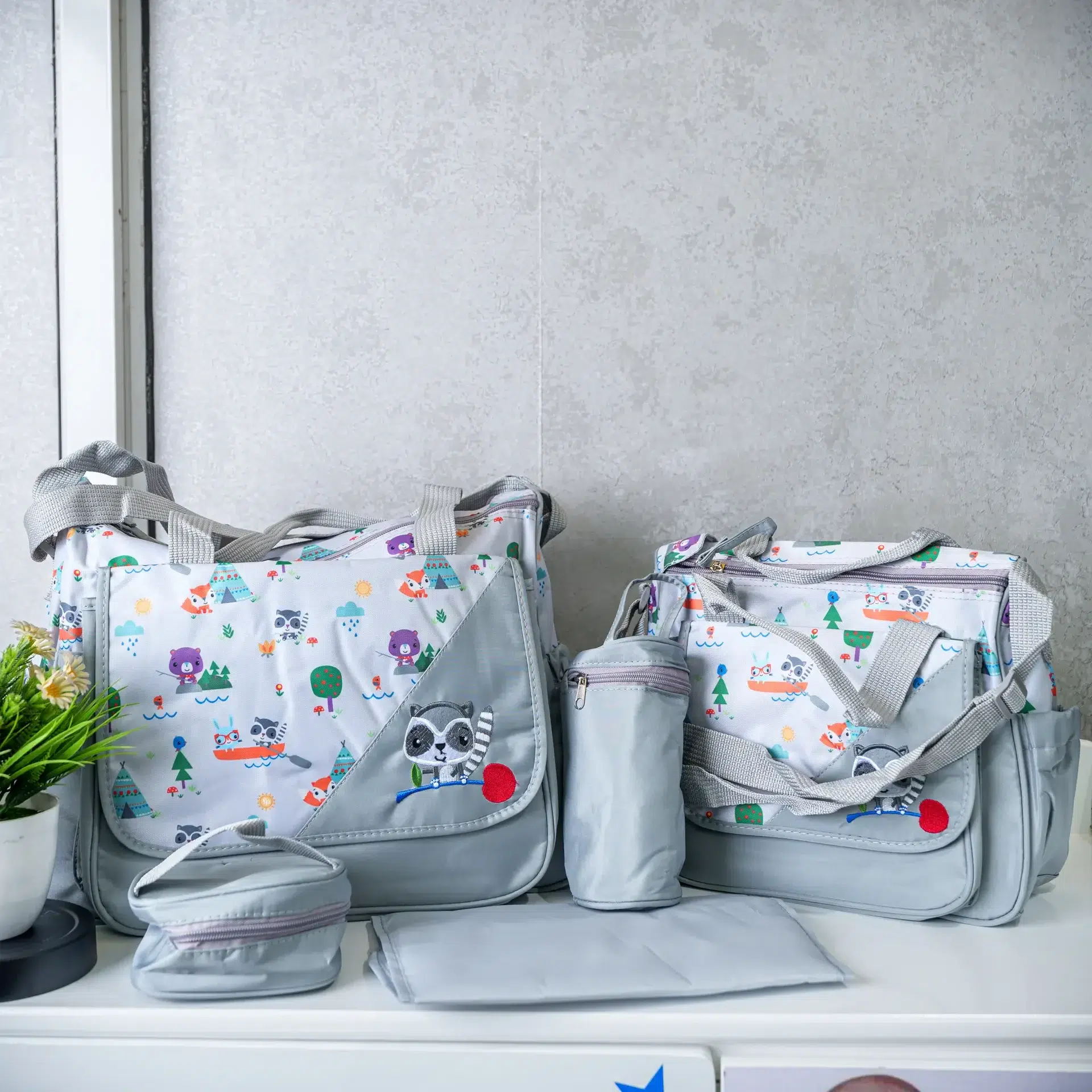 A five-piece diaper bag set displayed on a white surface, featuring two light gray bags with colorful cartoon animal prints, a matching bottle holder, a small pouch, and a foldable changing mat.