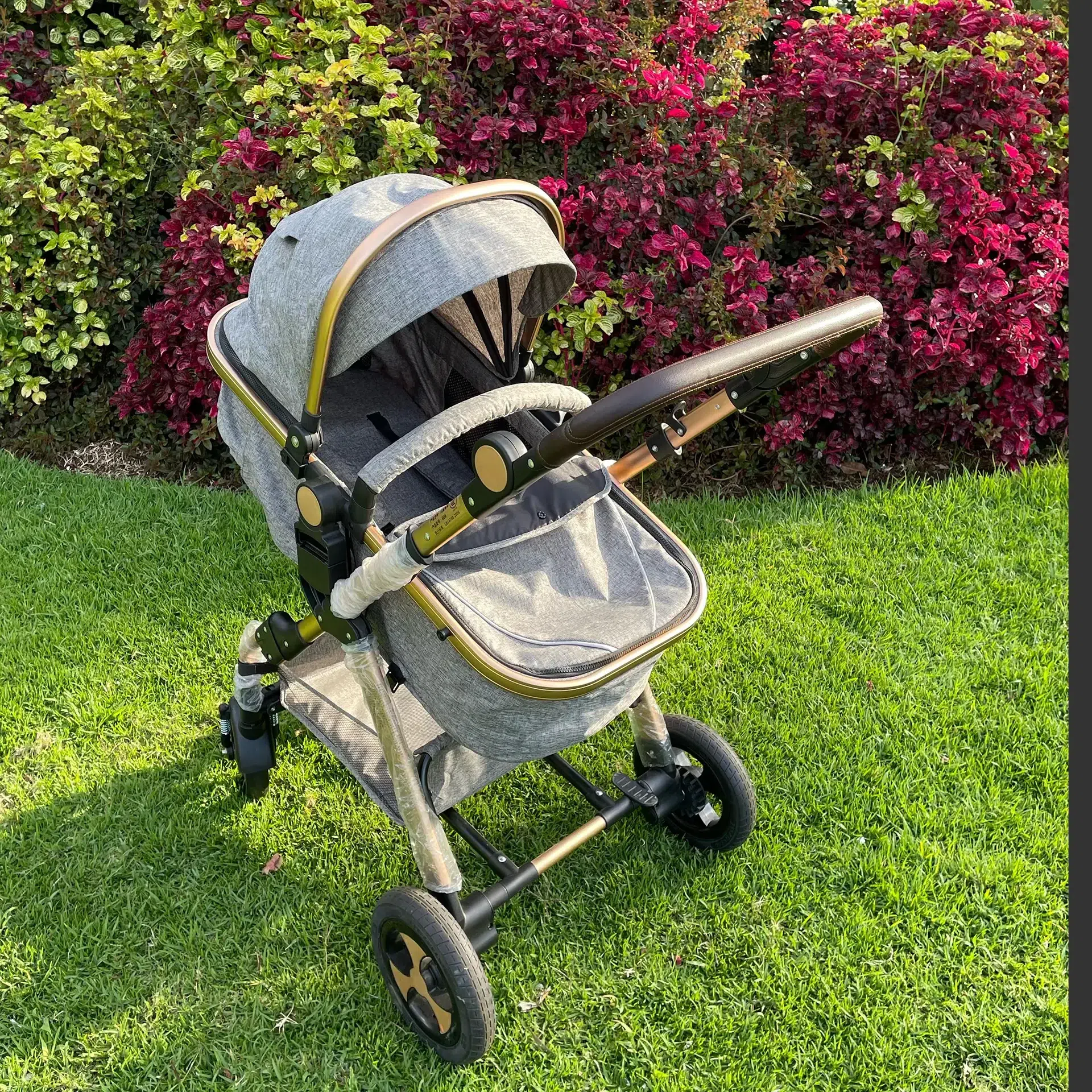Empty grey newborn bassinet stroller featuring a gold frame, adjustable canopy, large wheels, and under-seat storage basket, shown on grass.