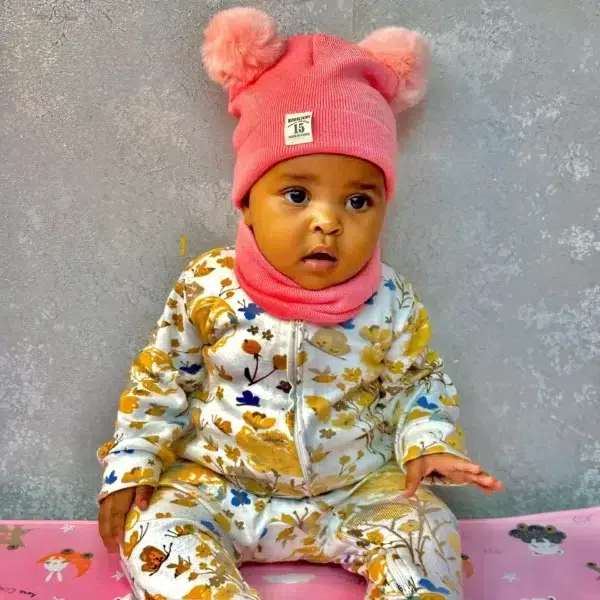 A baby wearing a pink beanie hat with two fluffy pom-poms and a matching neck warmer, dressed in a floral jumpsuit, sitting on a pink mat with cartoon prints.