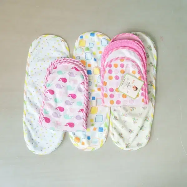 A collection of folded and unfolded baby burp cloths with playful designs, including whales, polka dots, stars, squares, and floral prints, arranged neatly on a flat surface.