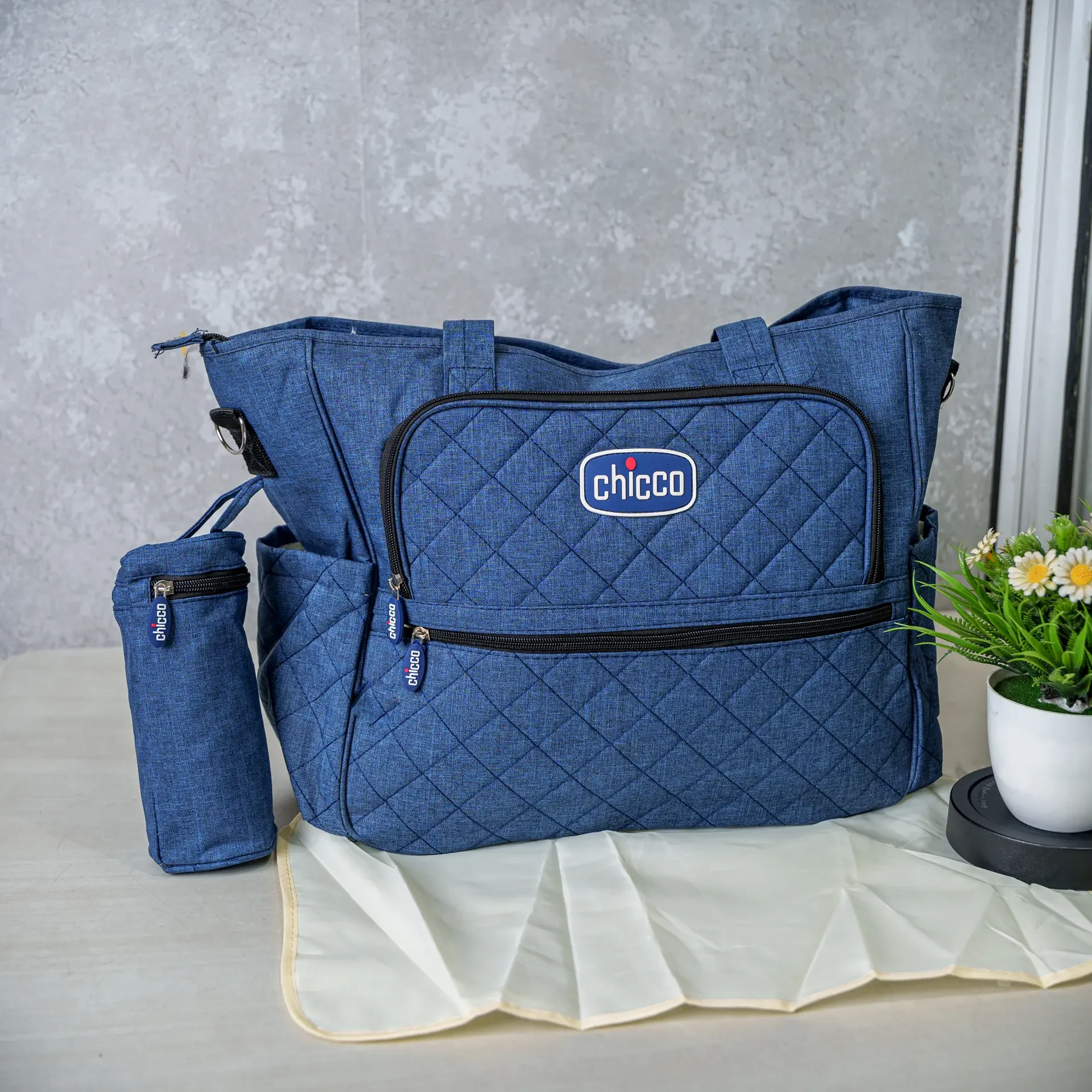A blue Chicco diaper bag set, including a quilted main bag with multiple compartments, a matching bottle holder, and a foldable changing mat, placed on a table next to a small decorative plant.