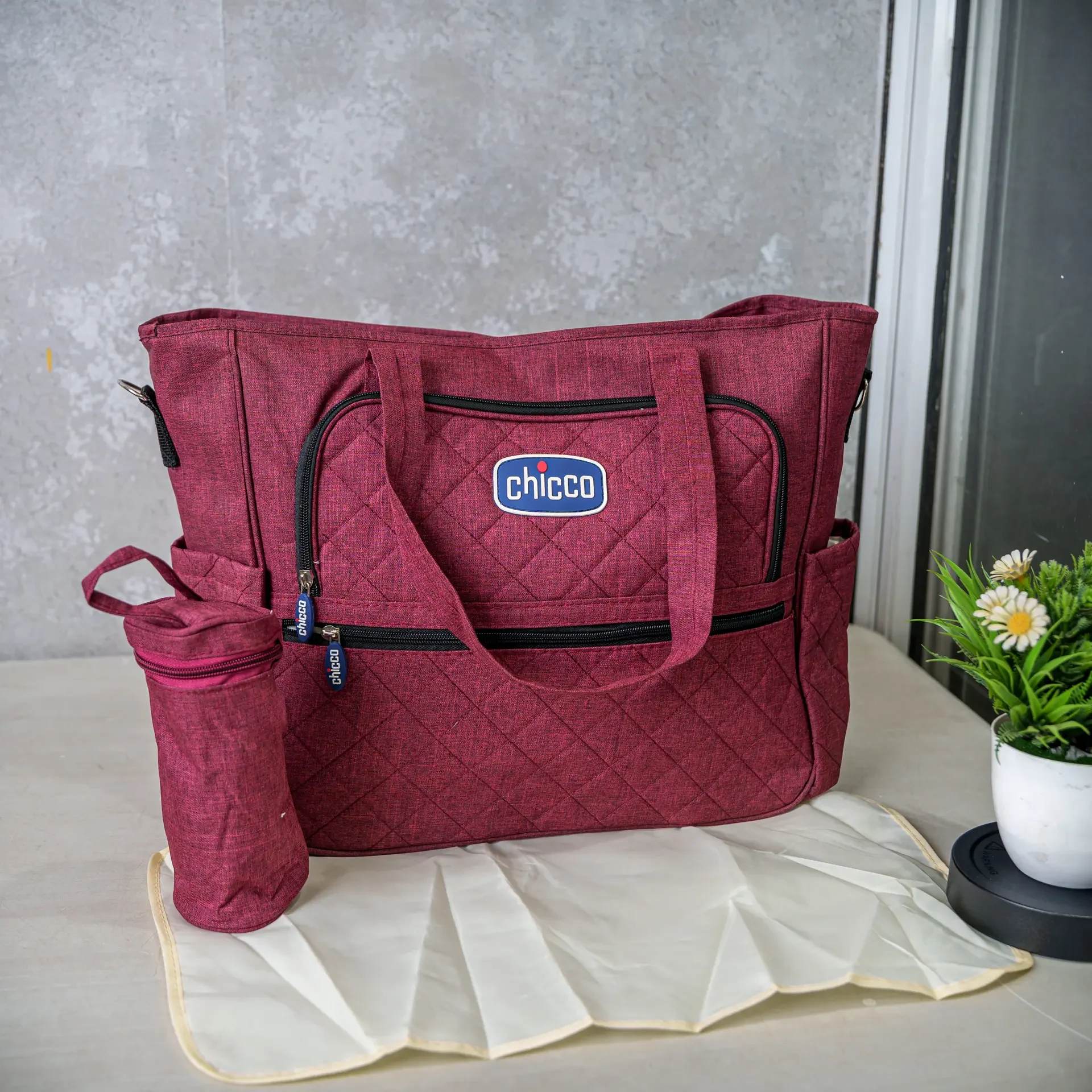 A red Chicco diaper bag set, featuring a quilted design, spacious compartments, a matching insulated bottle holder, and a foldable changing mat, displayed on a table with a decorative plant in the background.