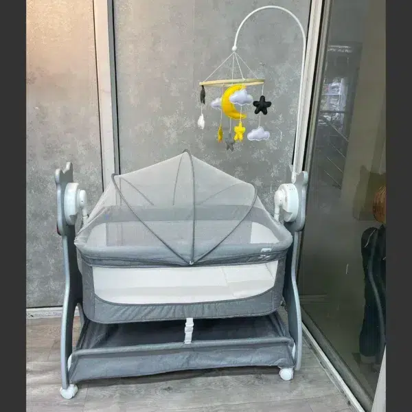 Modern grey electric rocking bassinet featuring breathable mesh sides, a protective canopy with mosquito net, a hanging moon and stars mobile, and a lower storage shelf on wheels.