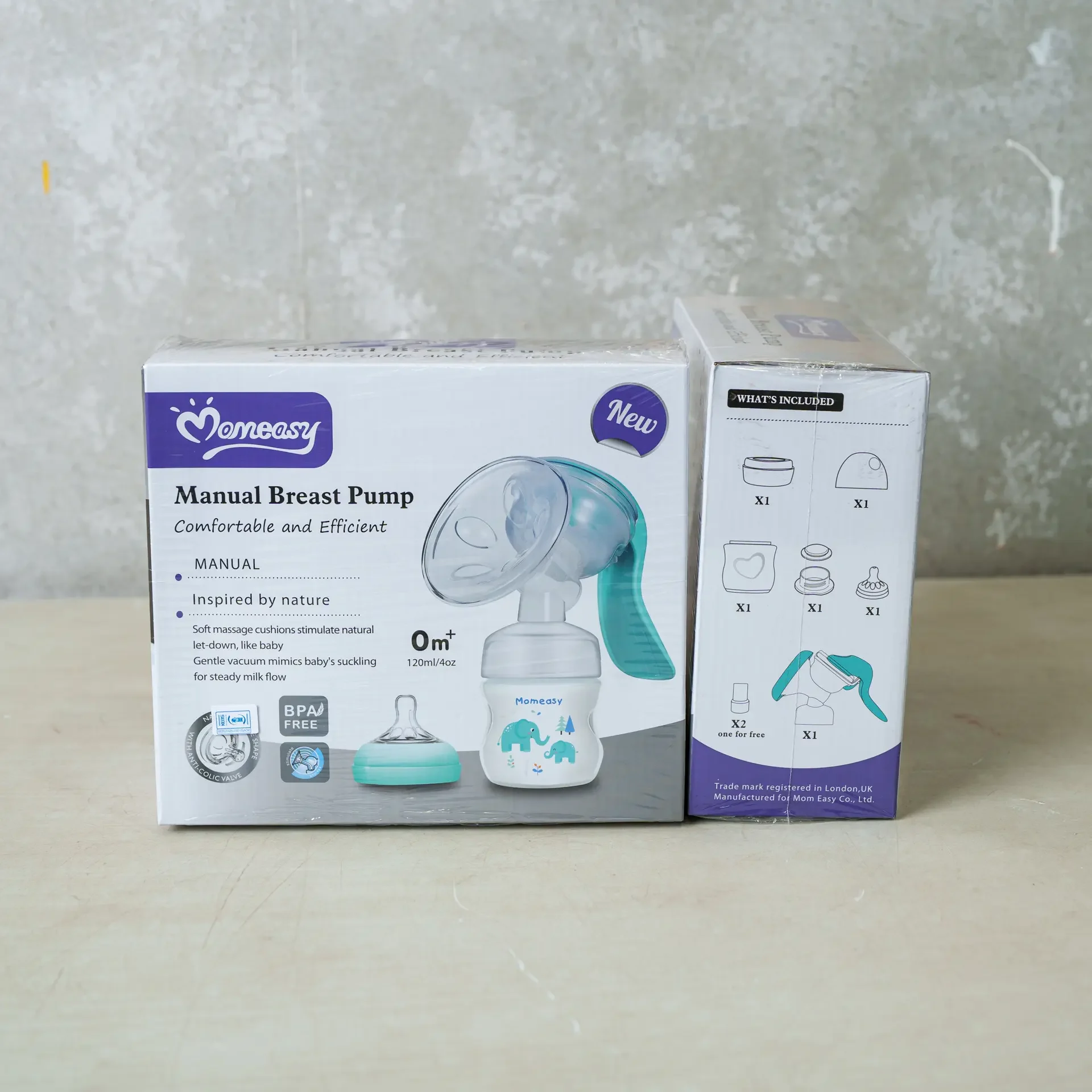 Box of Momeasy Manual Breast Pump placed on a light surface, showing the front and side packaging. The product includes a BPA-free 120ml/4oz bottle with a soft massage cushion and ergonomic handle, designed for comfortable and efficient milk expression. The side of the box lists included components such as the pump, bottle, nipple, cap, and accessories.