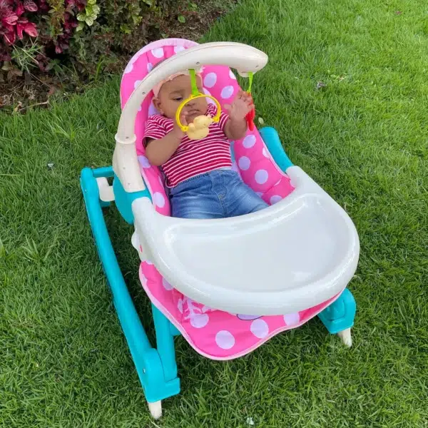 Baby girl sitting in the pink and turquoise 3-in-1 rocking chair/infant seat, playing with the attached toy bar outdoors.